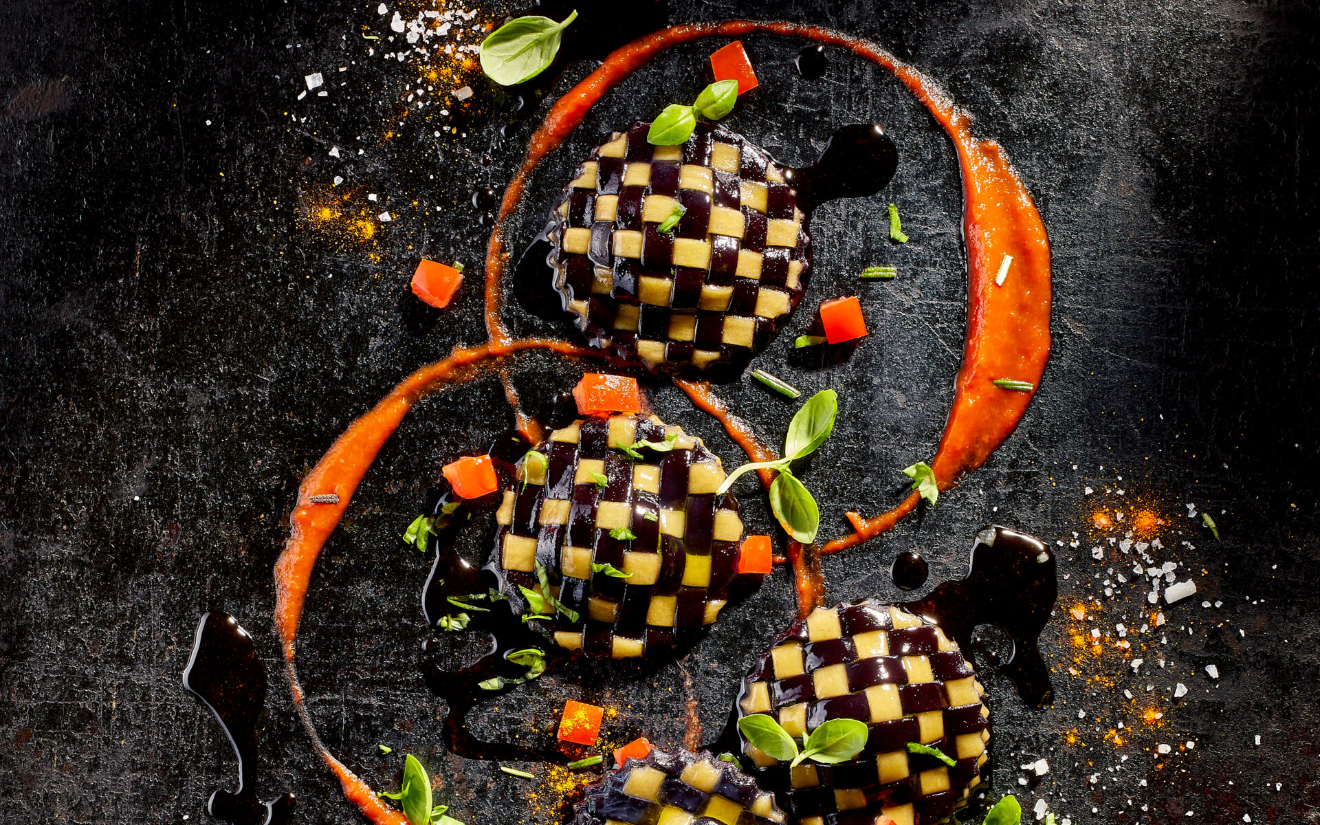 Drei Ravioli/ Pasta aus zweierlei Teig - dunklem und hellem - kunstvoll gewebt. Dekoriert mit Paprikastückchen, frischem Basilikum sowie Papikasauce. Auf schwarzem Untergrund elegant fotografiert.