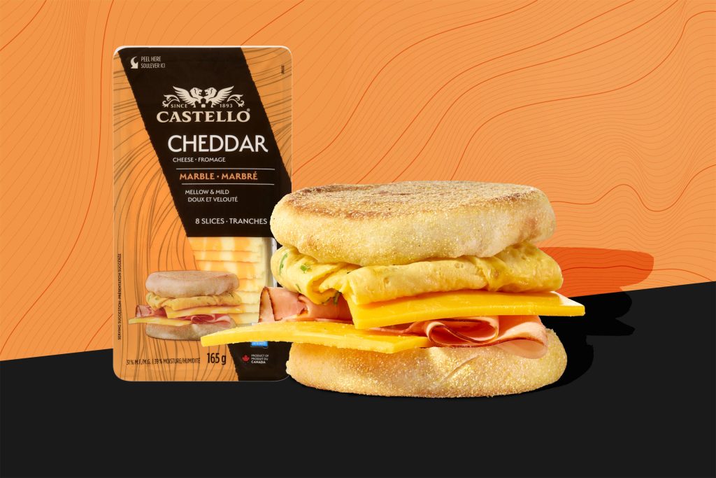 Castello Cheddar: gegrillter/ getoasteter English Muffin/ Frühstücksmuffin mit Rührei, gekochtem Schinken und Käse belegt. Auf schwarzem Grund mit Schattenwurf, im Hintergund eine orangene Fläche mit schwarzen Linien, als Motiv aufgenommen von der Cheddar-Verpackung, die daneben abgebildet ist.