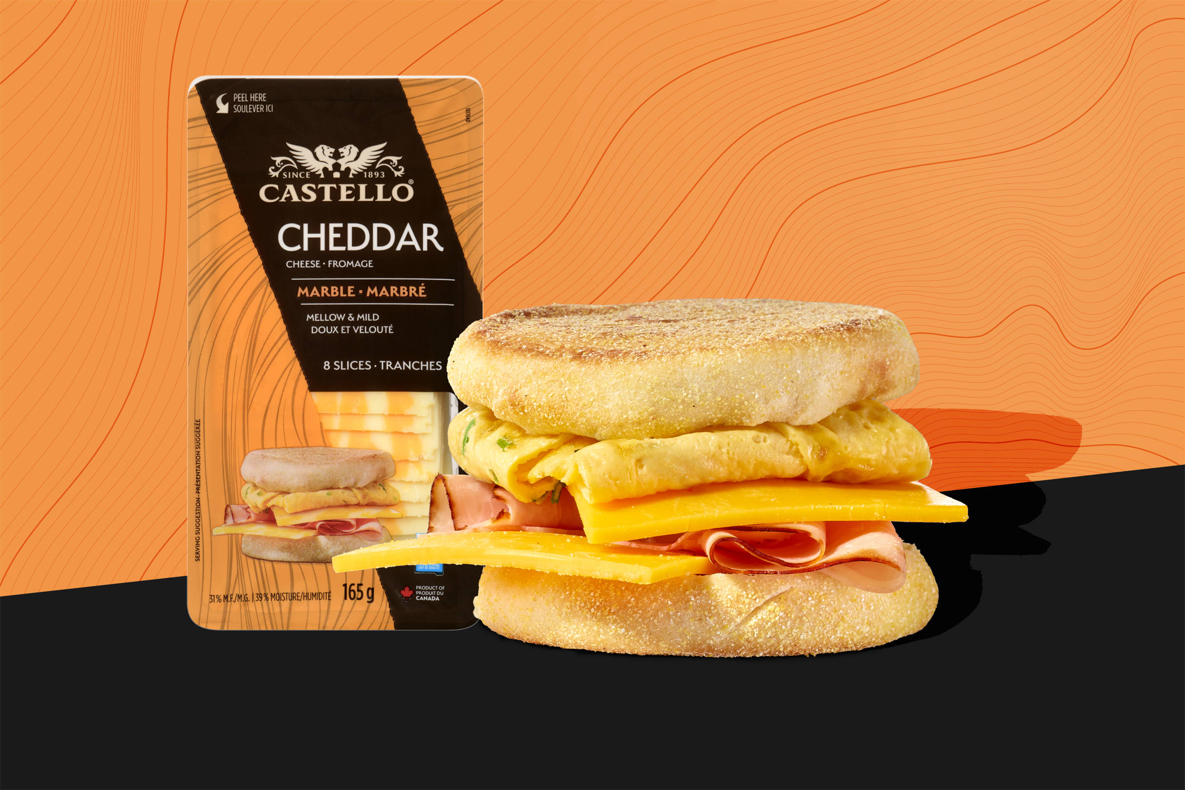 Castello Cheddar: gegrillter/ getoasteter English Muffin/ Frühstücksmuffin mit Rührei, gekochtem Schinken und Käse belegt. Auf schwarzem Grund mit Schattenwurf, im Hintergund eine orangene Fläche mit schwarzen Linien, als Motiv aufgenommen von der Cheddar-Verpackung, die daneben abgebildet ist.