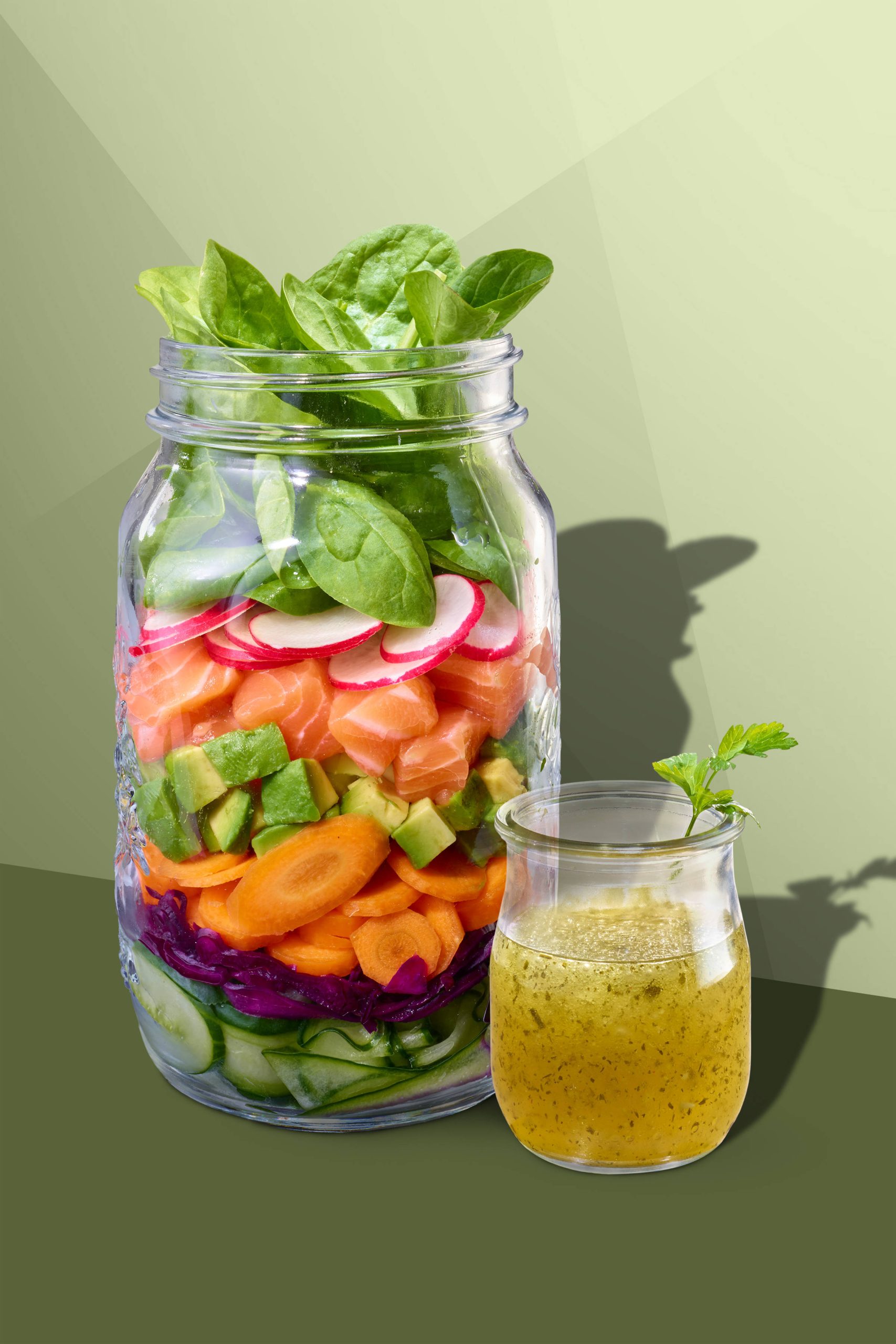 Großes Glas mit geschichtem Salatmix: Gurkenscheiben, eingelegte rote Zwiebel, Karottenscheiben, Avocadowürfel, frischer Lachs/ Lachs Sashimi, Radieschenscheiben und Spinatblätter, daneben ein Glas mit Salatsoße/ Vinaigrette. Auf dunkelgrünem Grund mit Schattenwurf.