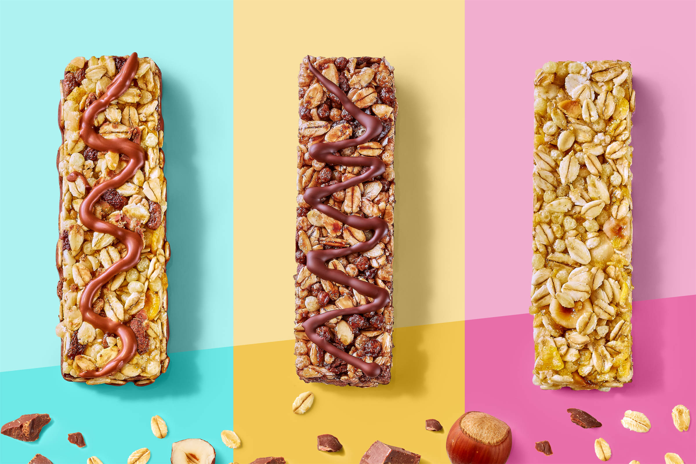 Drei Müsliriegel nebeneinander auf je unterschiedlichen Farbflächen: links türkis (Schoko-Banane), Mitte gelb (Schokolade), rechts rosa (Haselnuss). Dekoriert mit Nüssen, Haferflocken und Schokokrümeln.