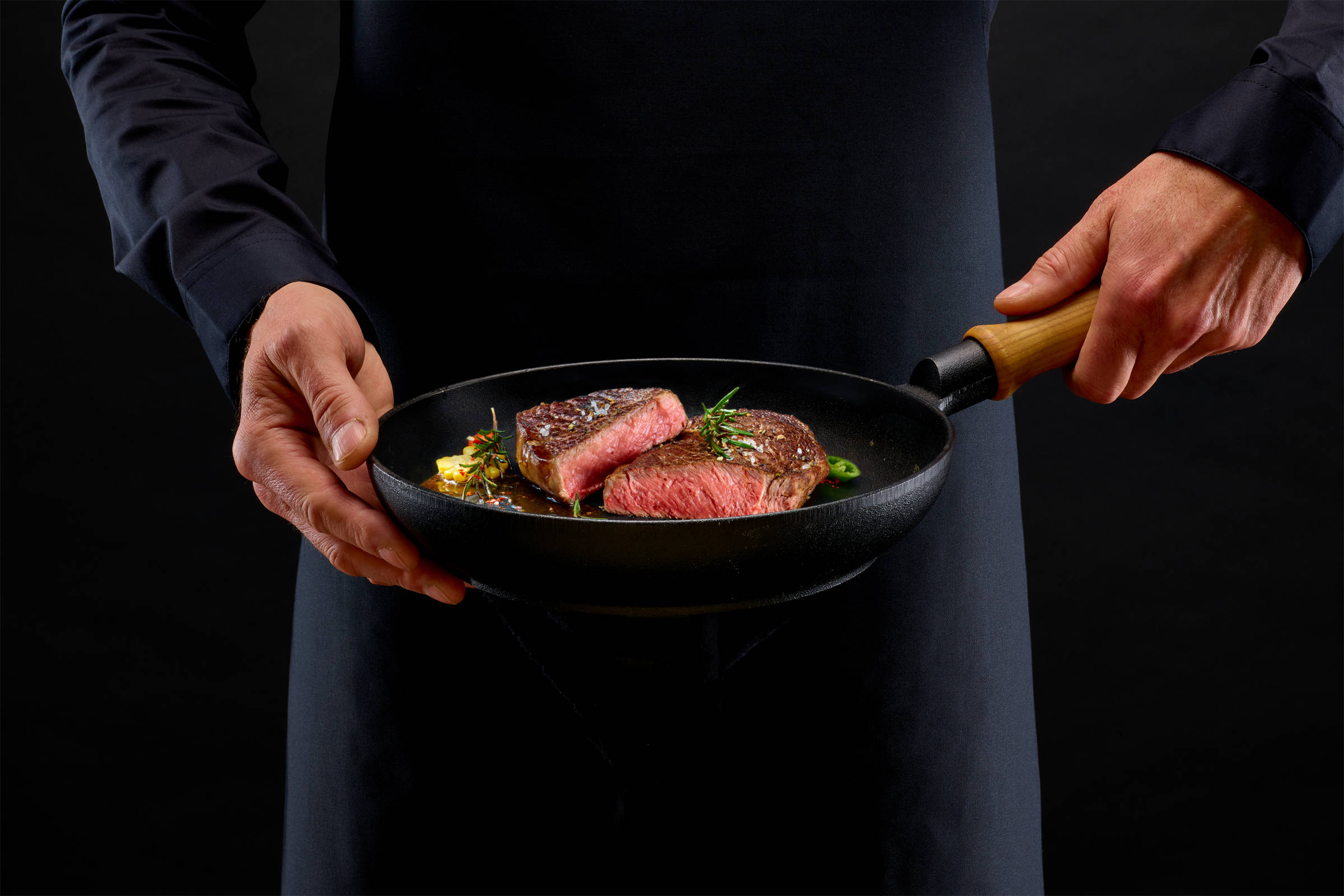 Zwei Männerhände halten eine gusseiserne Pfanne mit einem gebratenen Rumpsteak/ Steak, medium rare. Darauf Rosmarin. Vor schwarzem Hintergrund.