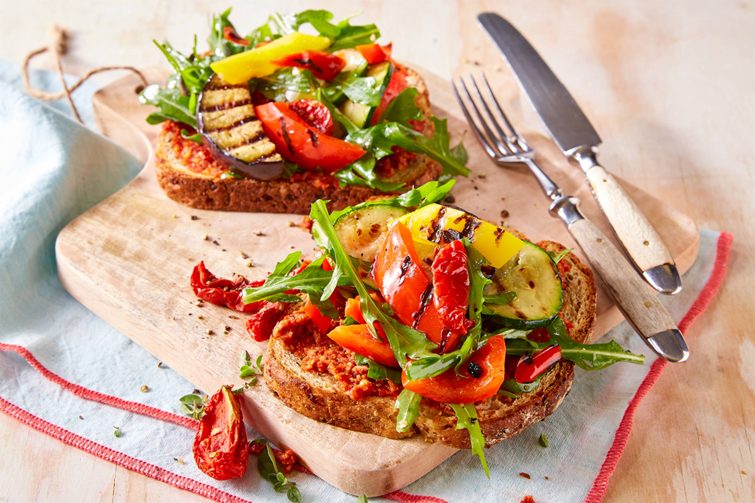 2 Zwei Toast mit Tomaten, gegrillter Paprika, Aubergine & Zucchini, Rukola auf einem Holzbrett mit Messer & Gabel.