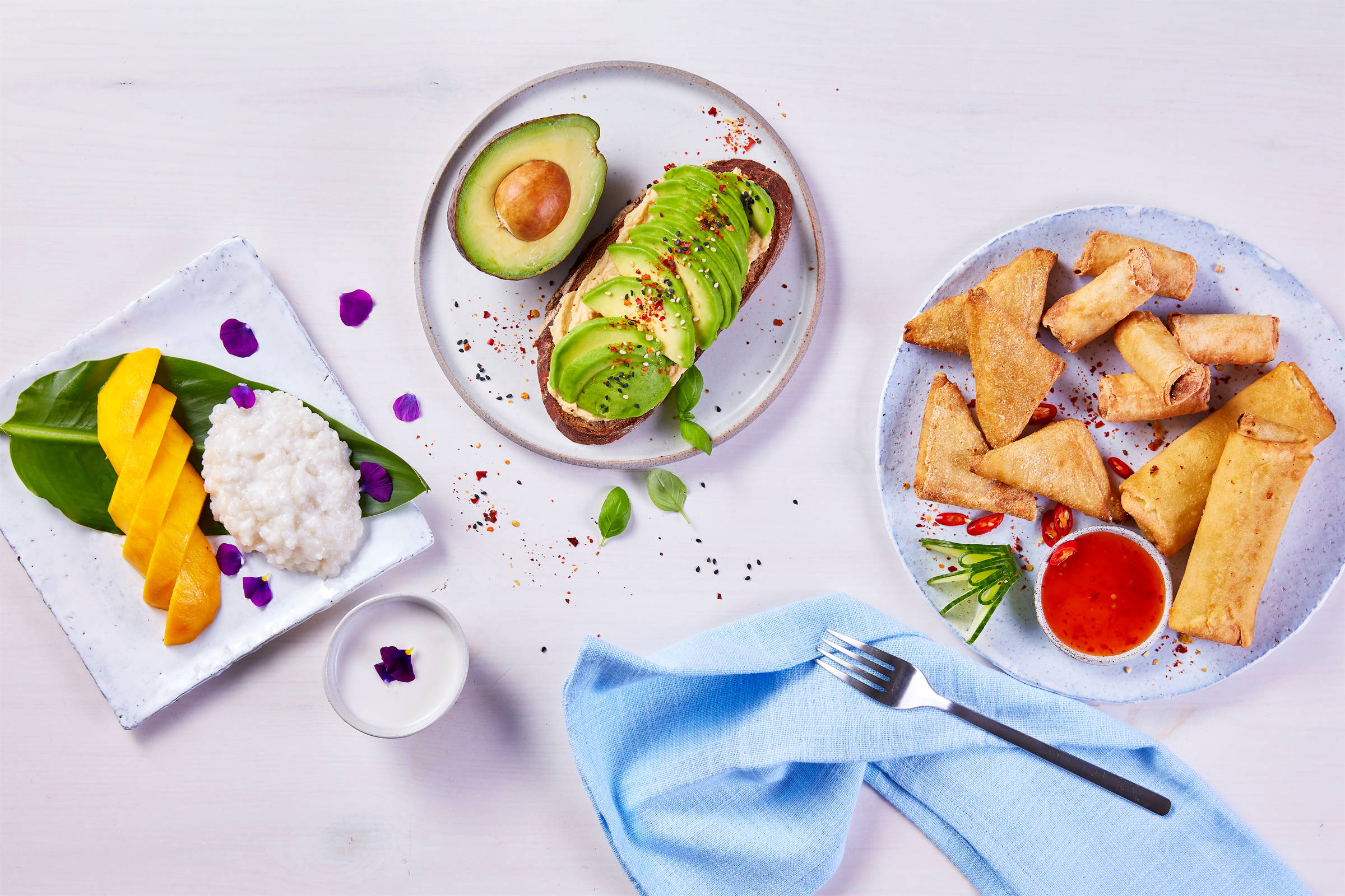 Paulsen: links ein Teller Reis und Mangostücken, mittig ein Teller mit einem Brotmit Avocadoscheiben belegt; rechts ein Teller mit Frühlingsrollen, Pakoras und roter Eintunksoße. Und eine blaue Stoffserviette sowie eine Gabel.