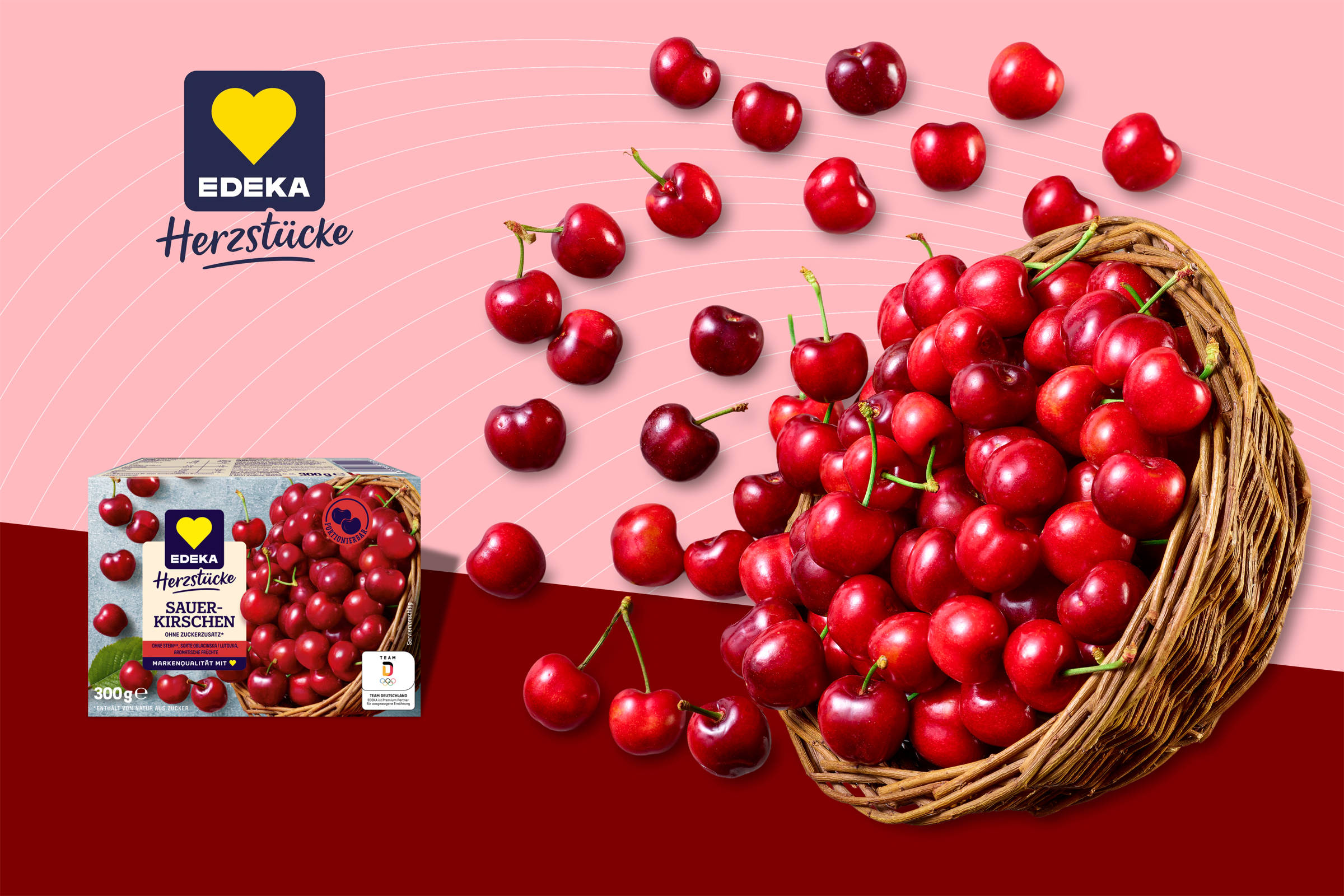 Edeka Herzstücke: Aus einem geflochteten Korb fallen viele Kirschen vor einem zweigeteilten Hintergrund. Oben rosa mit dünnen weißen kreisförmigen Streifen, unten ein dunkles Weinrot. Links im Bild die Verpackung.
