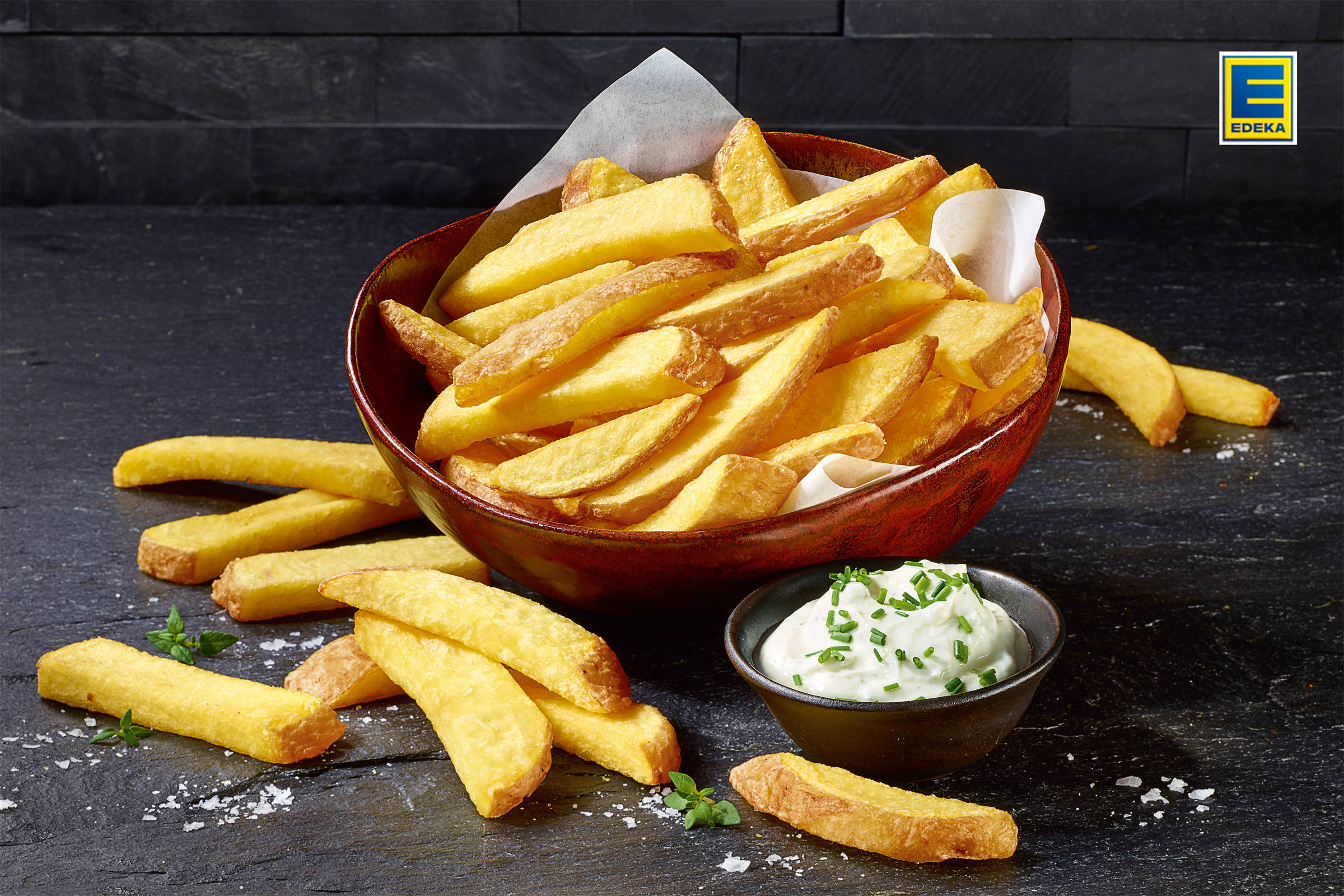 Edeka: Pommes Frites in einer Schale auf schwarzem Schiefer, daneben kleine Schale mit Sour Cream/ Schnittlauch.