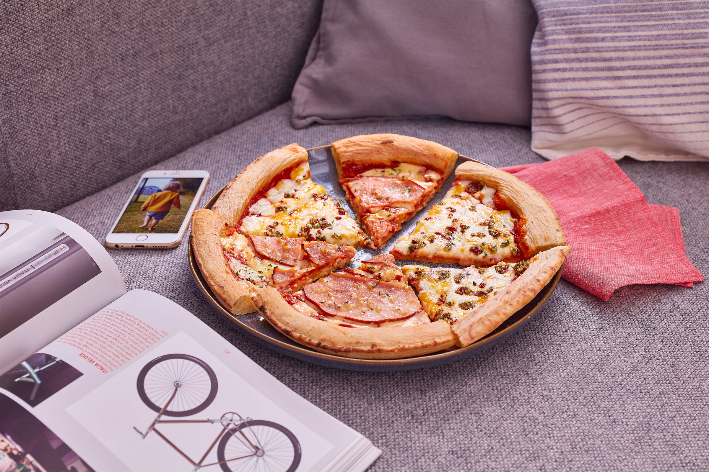 Eine zerteilte Pizza auf einem Teller, der auf einem grauen Sofa liegt. Daneben ein Fahrradkatalog, ein Handy mit dem Foto eines Kindes und eine rote Stoffserviette.