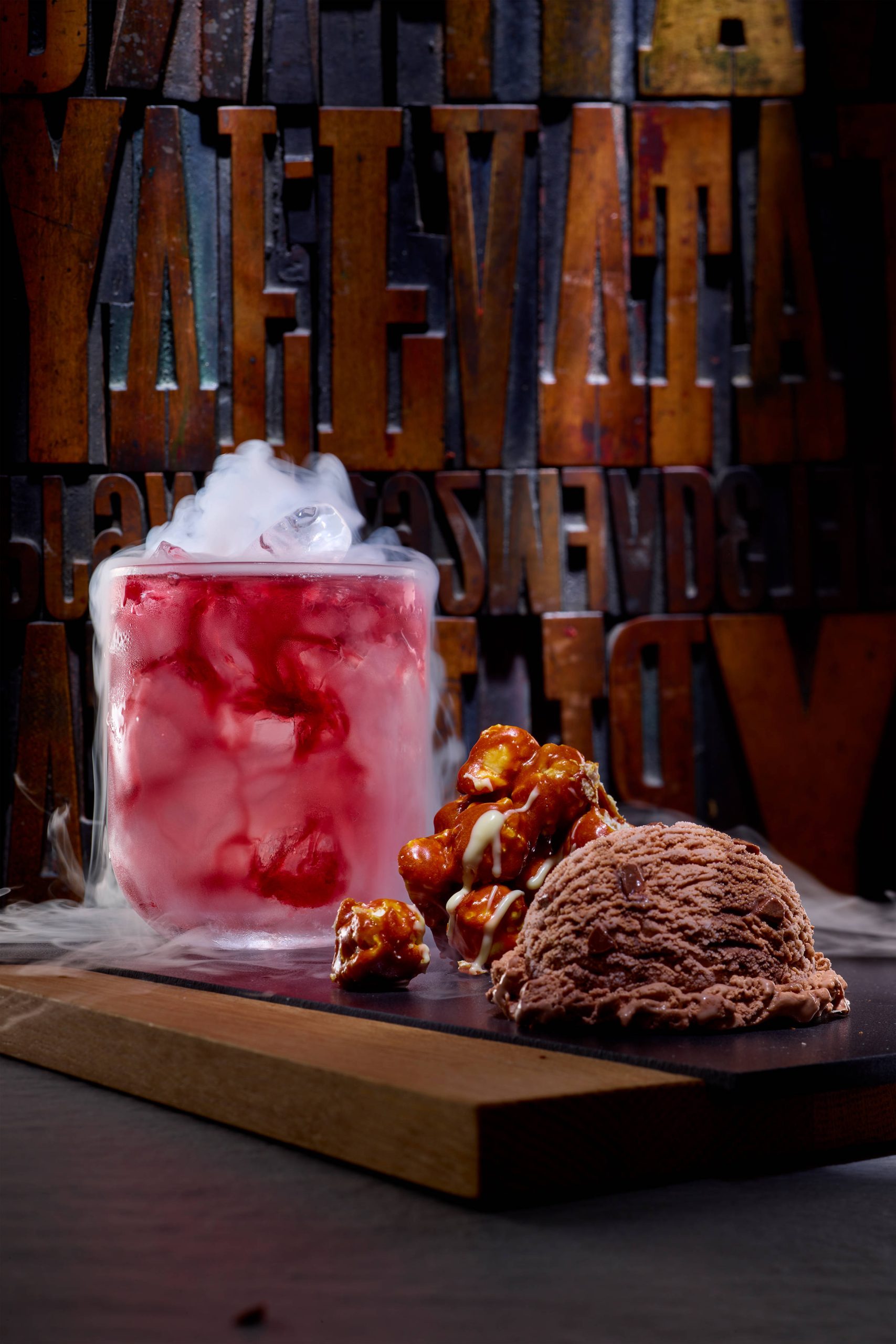 Auf einem Holzbrett liegt eine Kugel Schokoladeneis, daneben etwas Karamelisiertes, dahinter ein Glas mit vielen Eiswürfeln und einer roten Flüssigkeit im Eisnebel.