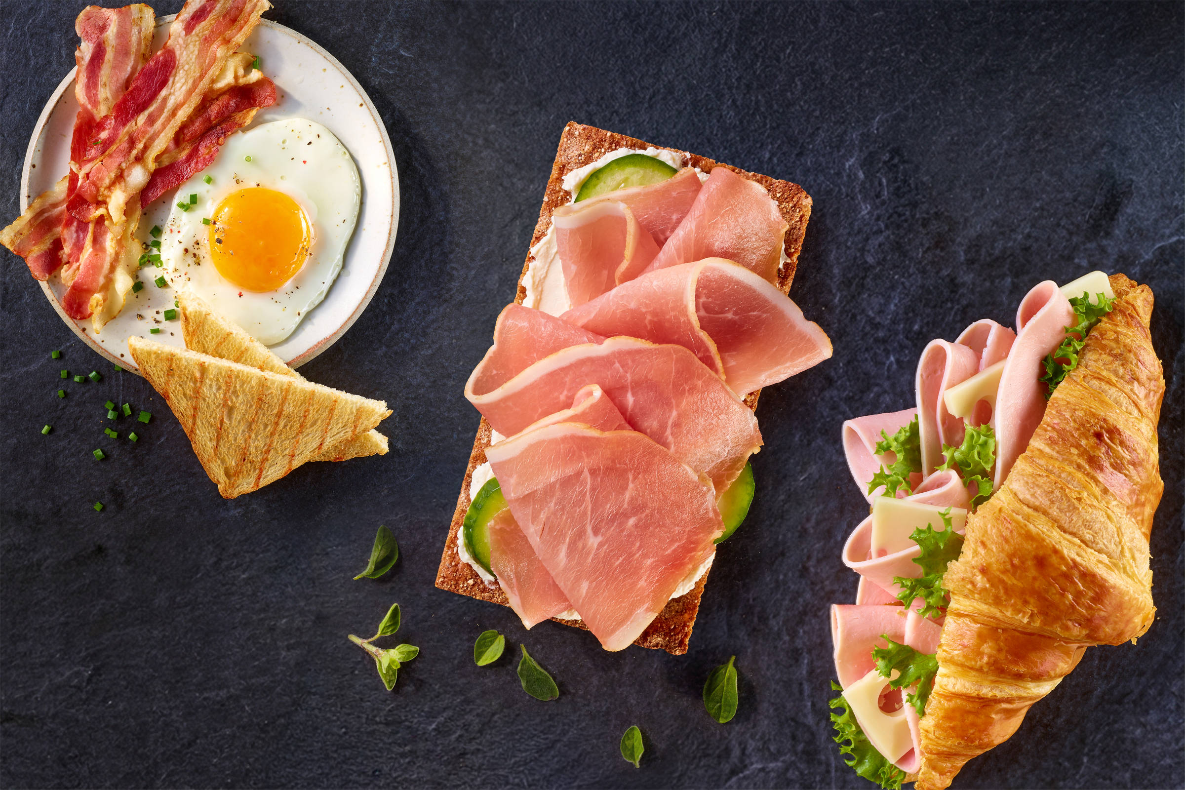 Ein Teller mit Spiegelei, Bacon und Toast, 1 Knäckebrot mit Frischkäse, Gurkenscheiben und Schinken, 1 Croissant mit Leerdamer Käse, Wurstscheiben und grünem Salat gefüllt. Alles auf schwarzem Schiefer von oben aufgenommen.