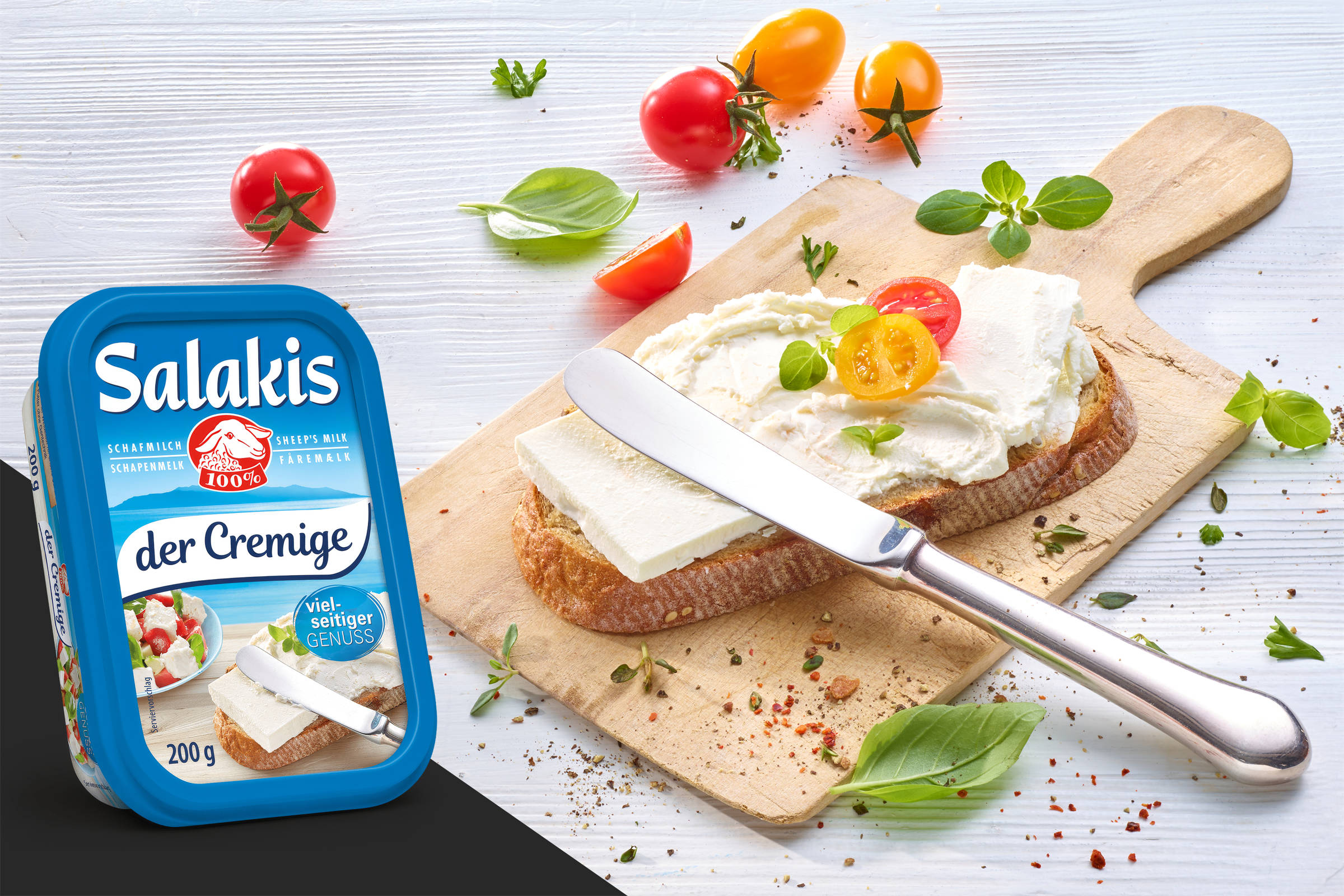 Salakis Frischkäse aus Schafmilch: Ein Holzbrettchen mit einer mit Frischkäse geschmierten Scheibe Brot, dazu Kirschtomaten, Basilikumblätter und ein Messer. Die Verpackung ist ebenfalls abgelichtet.