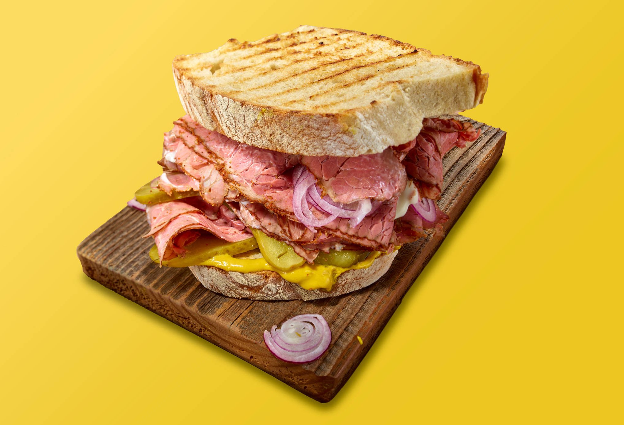 Ein getoastetes Sandwich mit Roastbeef, Gurkenscheiben und Senfsauce auf einem groben Holzbrett vor einfarbig gelbem Hintergrund aufgenommen. Schräger, diagonaler Anschnitt.
