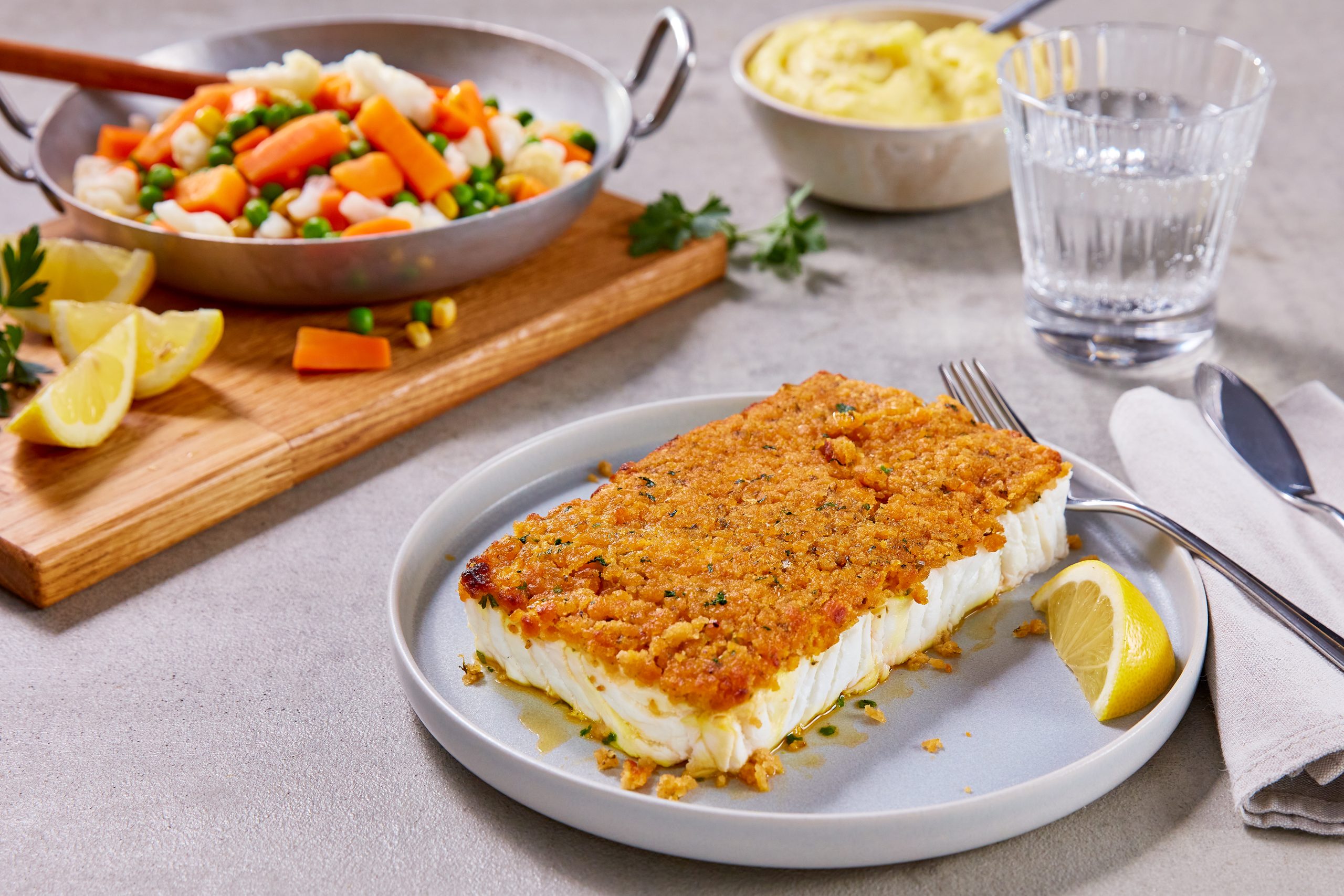 Frosta Schlemmerfilet à la Bordelaise Fischfilet auf weißem Teller auf gedecktem Tisch, daneben ein Glas Wasser, Kartoffelpürree und eine Pfanne mit Gemüse.